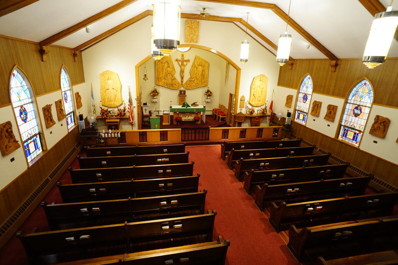 View of church from balcony with lines of wooden pews separated by a center aisle leading to the front altar
